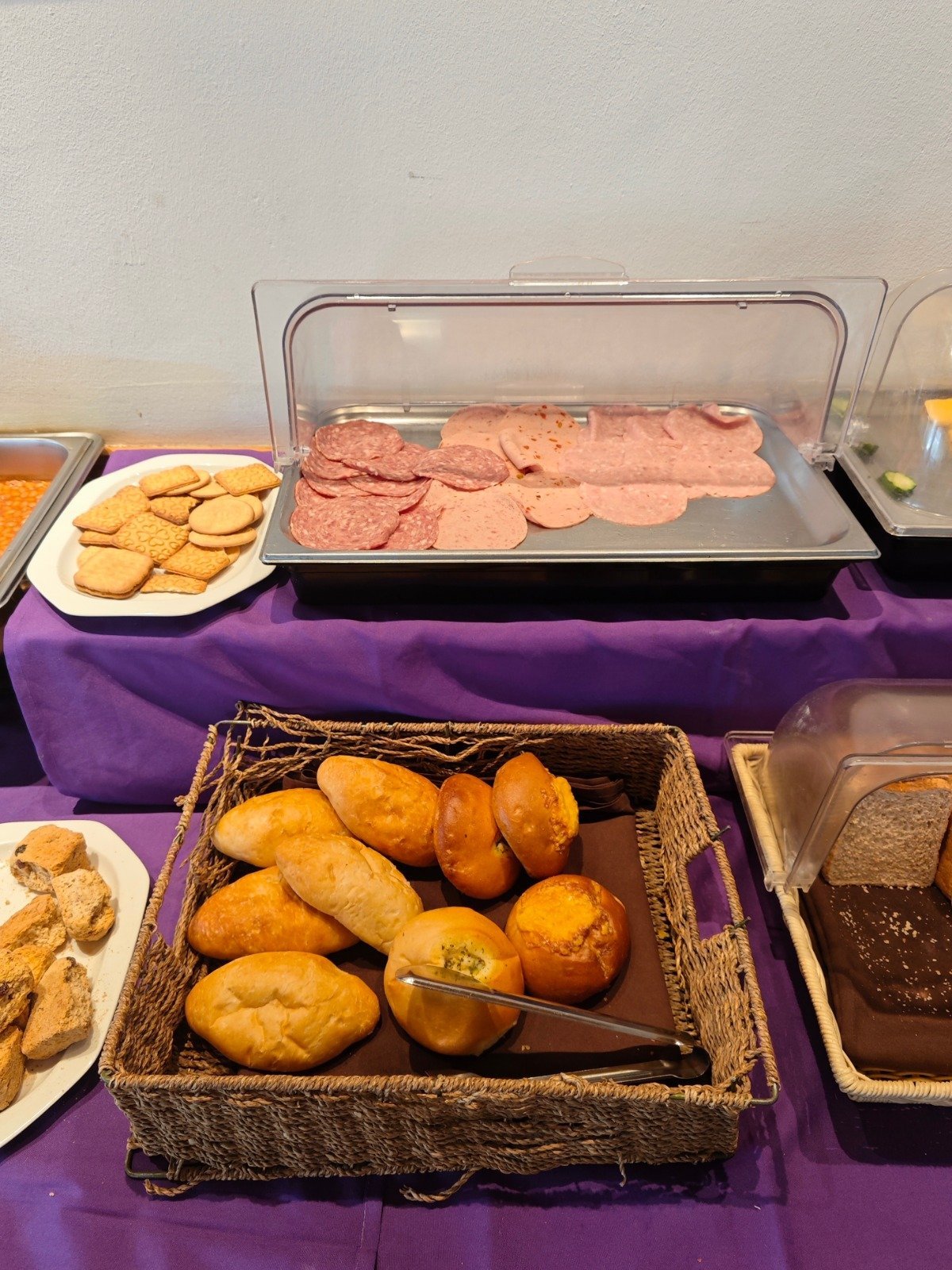 Buffet spread with fried bread rolls in a basket, sliced cured meat in a clear container, and cookies on a purple tablecloth