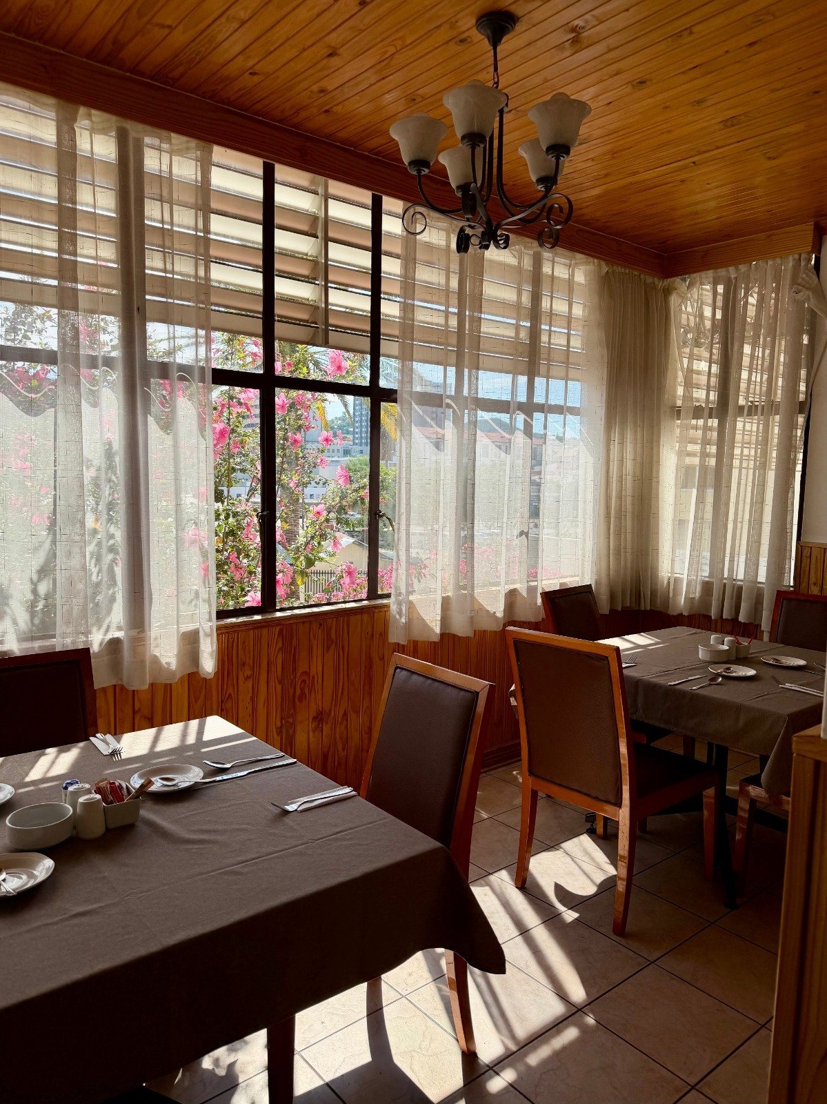 Bright dining room with wood ceiling, multiple windows with sheer curtains overlooking flowering garden, wooden furniture, and pendant chandelier