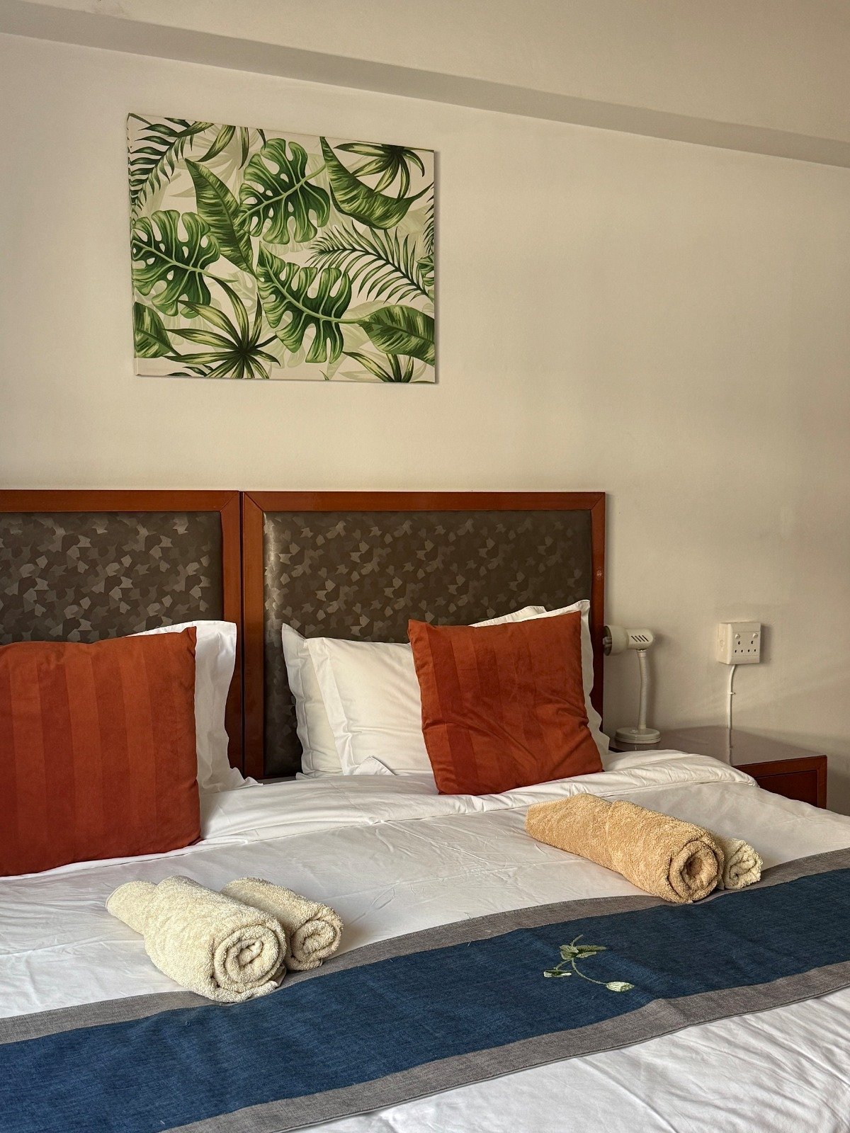 Hotel bedroom with wooden headboard, white bedding, red pillows, rolled towels, and tropical leaf artwork above
