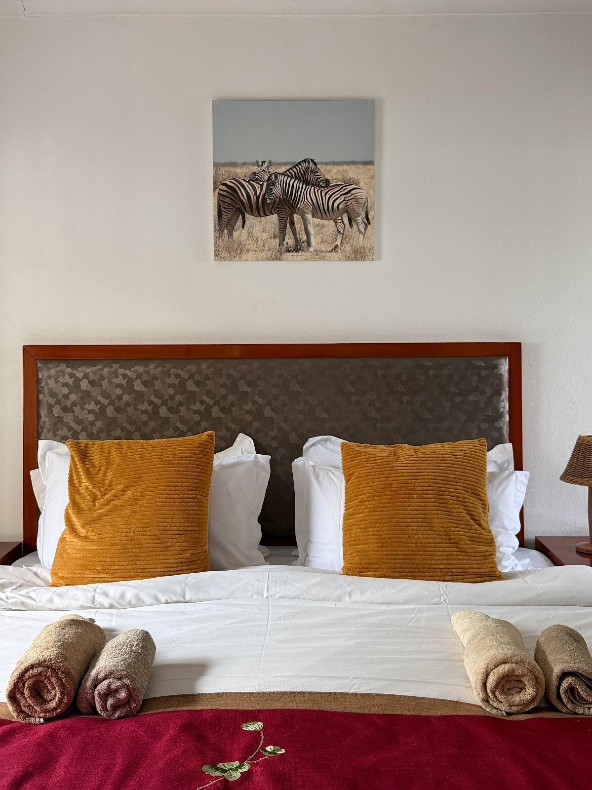 Modern bedroom with wooden bed frame, gray headboard, white pillows, golden cushions, rolled towels, and zebra wildlife artwork on wall