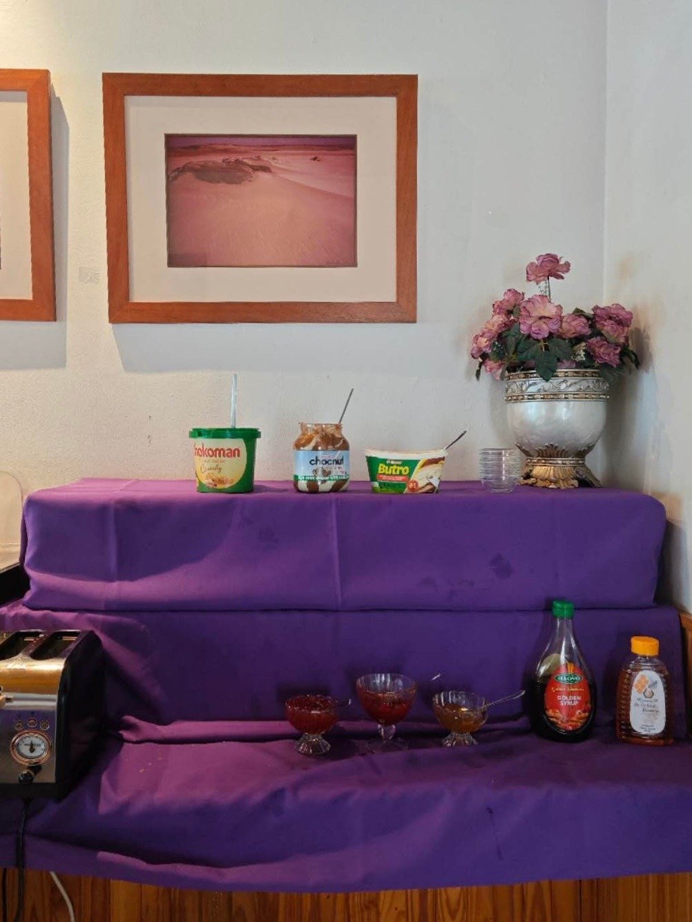 Purple sofa with framed pink landscape art on wall above, decorated with potted pink flowers and food containers on top