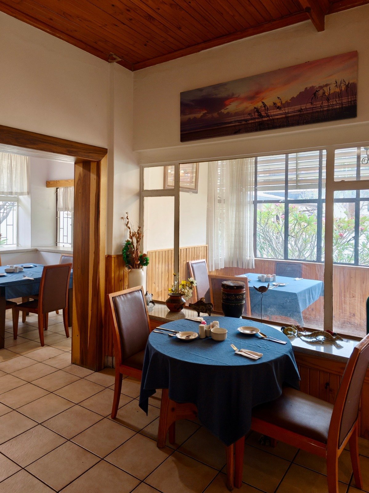 Elegant dining room with round blue tablecloth table, wooden chairs, large windows with curtains, wooden ceiling, and sunset artwork on wall