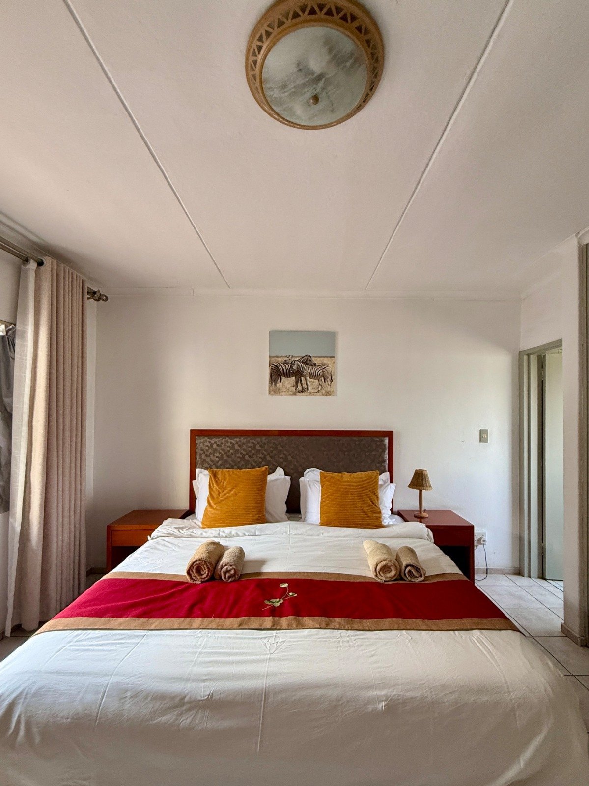 Modern bedroom with white walls, wooden bed frame, mustard and gold pillows, red accent runner, and a decorative ceiling clock