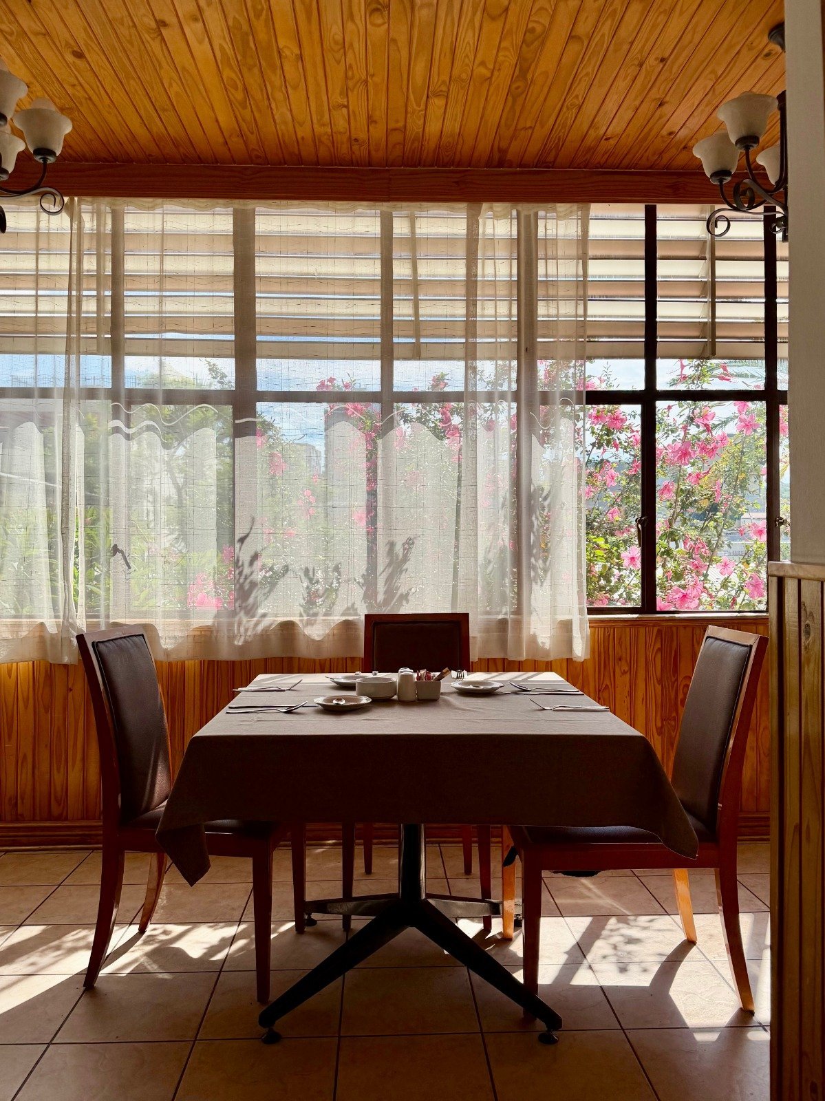 A dining room with a wooden ceiling, set table with two chairs, sheer curtains, and a view of flowering bushes outside through large windows.