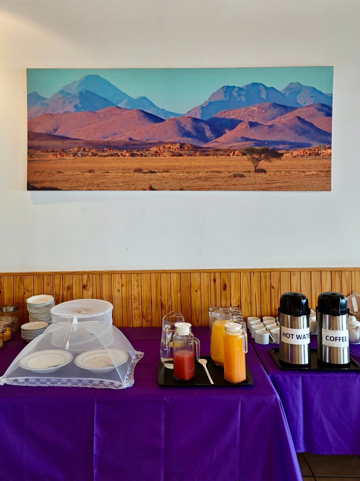 Breakfast buffet setup with purple tablecloth, plates, beverages, and desert mountain landscape photograph on wall
