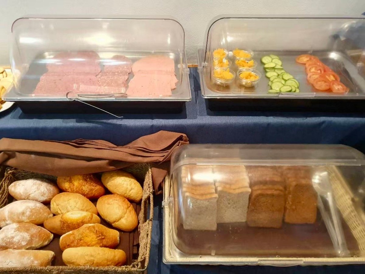 Buffet display with clear containers holding sliced deli meats, vegetables, rolls, and sauces on a dark counter
