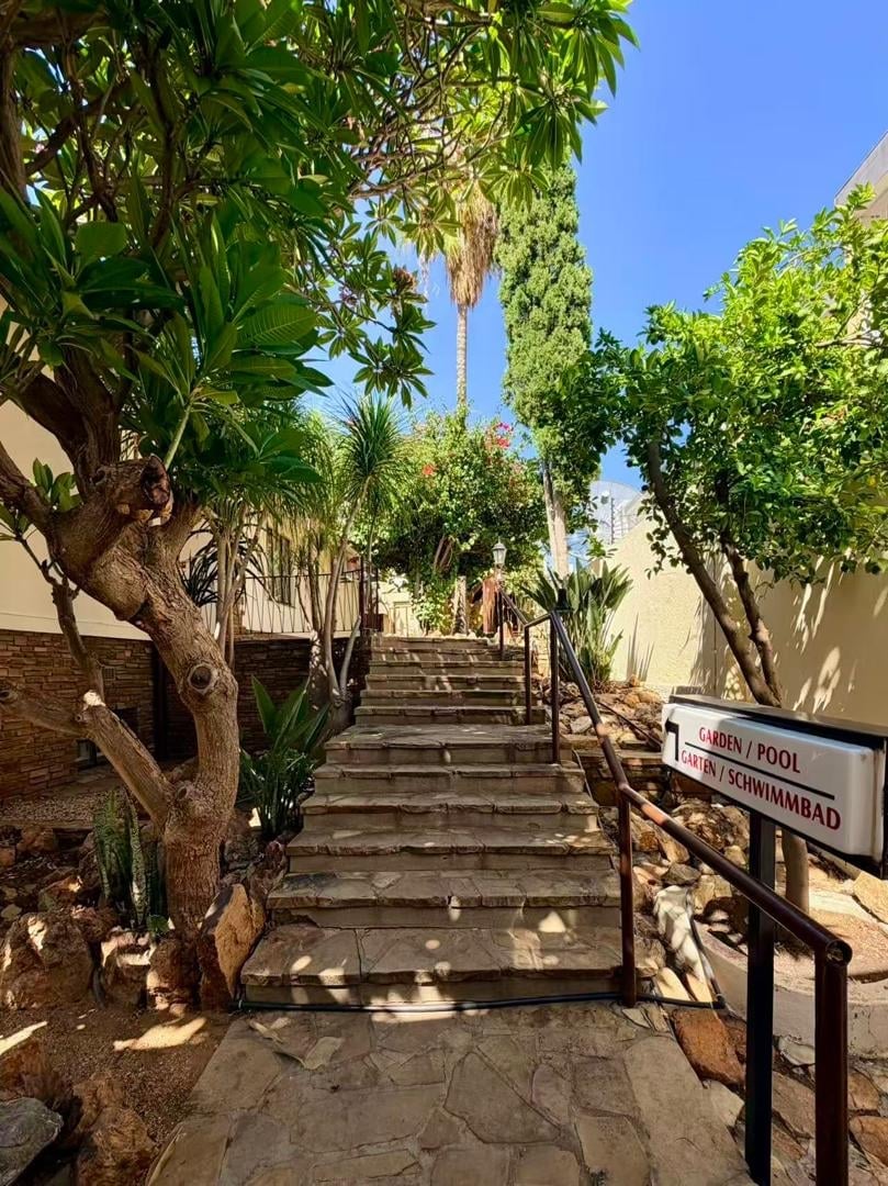 A shaded stone pathway lined with palm trees and tropical plants leading uphill toward a building under blue sky
