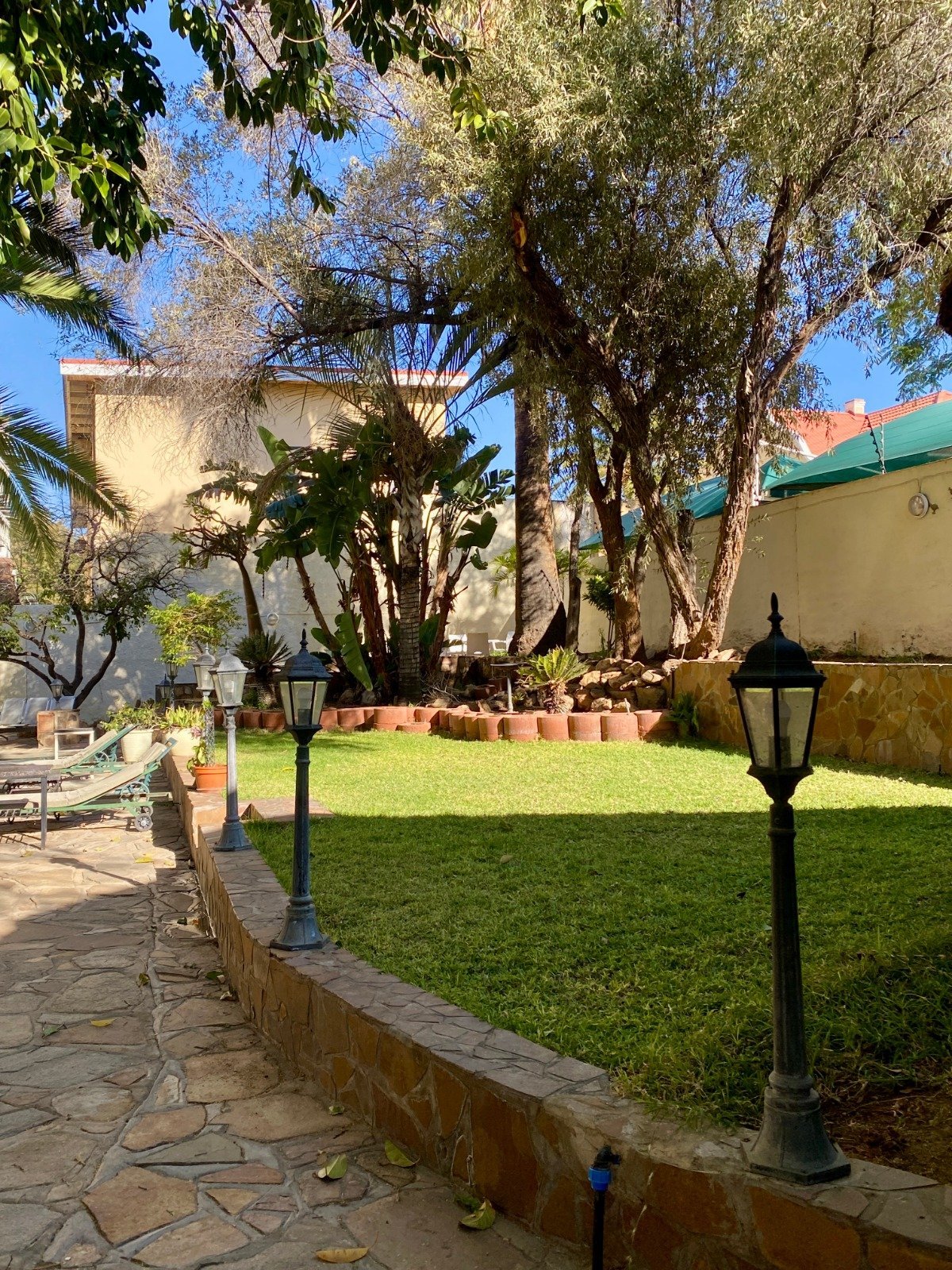 Sunny courtyard with green lawn, mature trees, historic stone building, and black lamp post along stone pathway