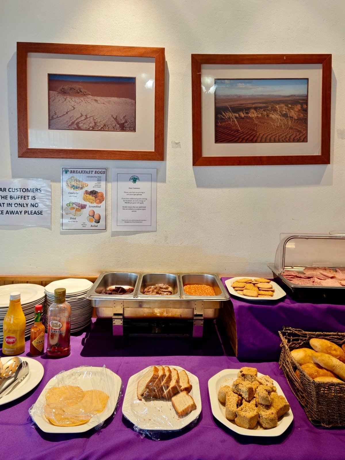 Buffet spread with framed desert landscape photos on white wall, purple tablecloth with bread, pastries, and food service trays below