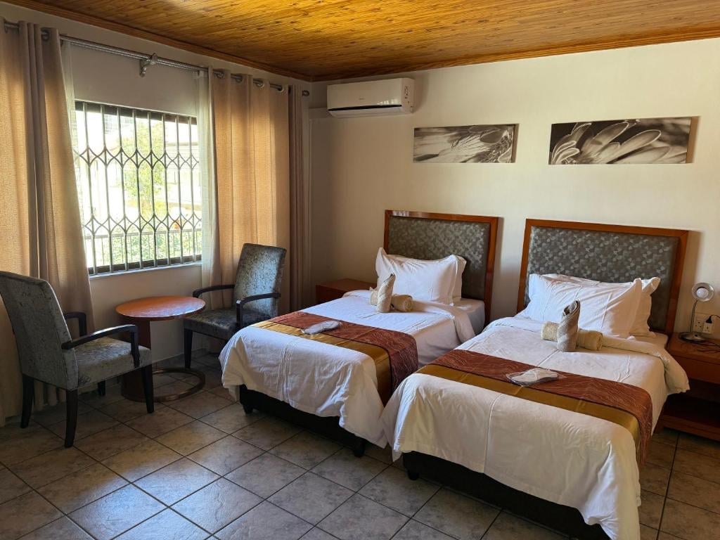 Hotel room with two twin beds, wooden ceiling, sitting area by window with terracotta curtains, and wall art