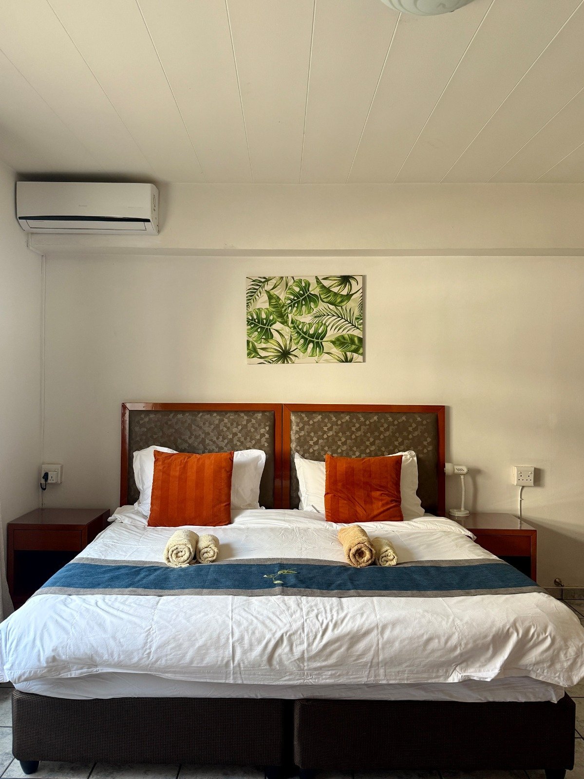 Modern bedroom with wooden bed frame, white bedding, orange pillows, botanical artwork, and air conditioning unit on cream walls