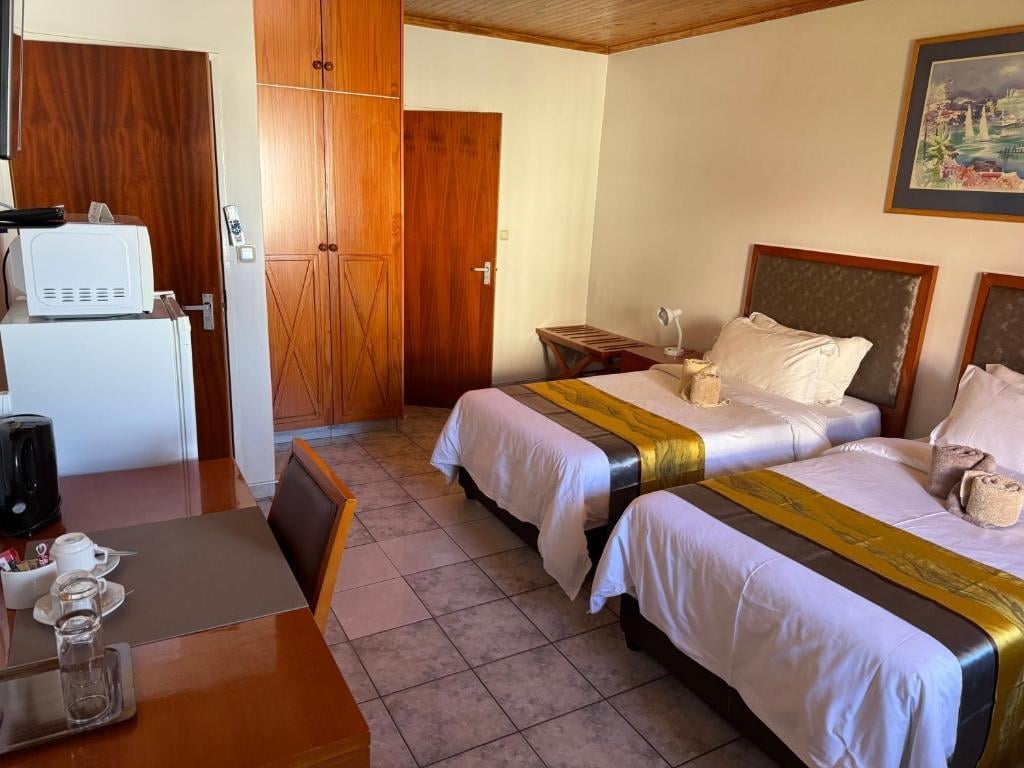 Hotel room with two twin beds with white linens and gold runners, wooden furniture, wardrobe, and mini-fridge