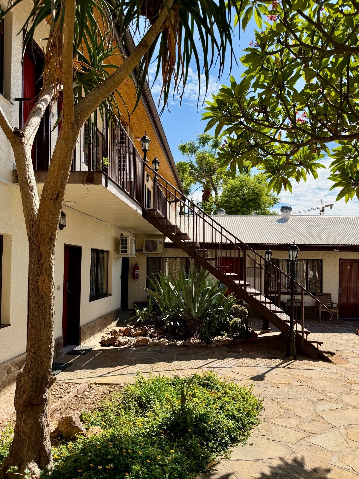 Courtyard of a colonial-style building with white and cream walls, wooden staircase, palm trees, and lush green vegetation under blue sky