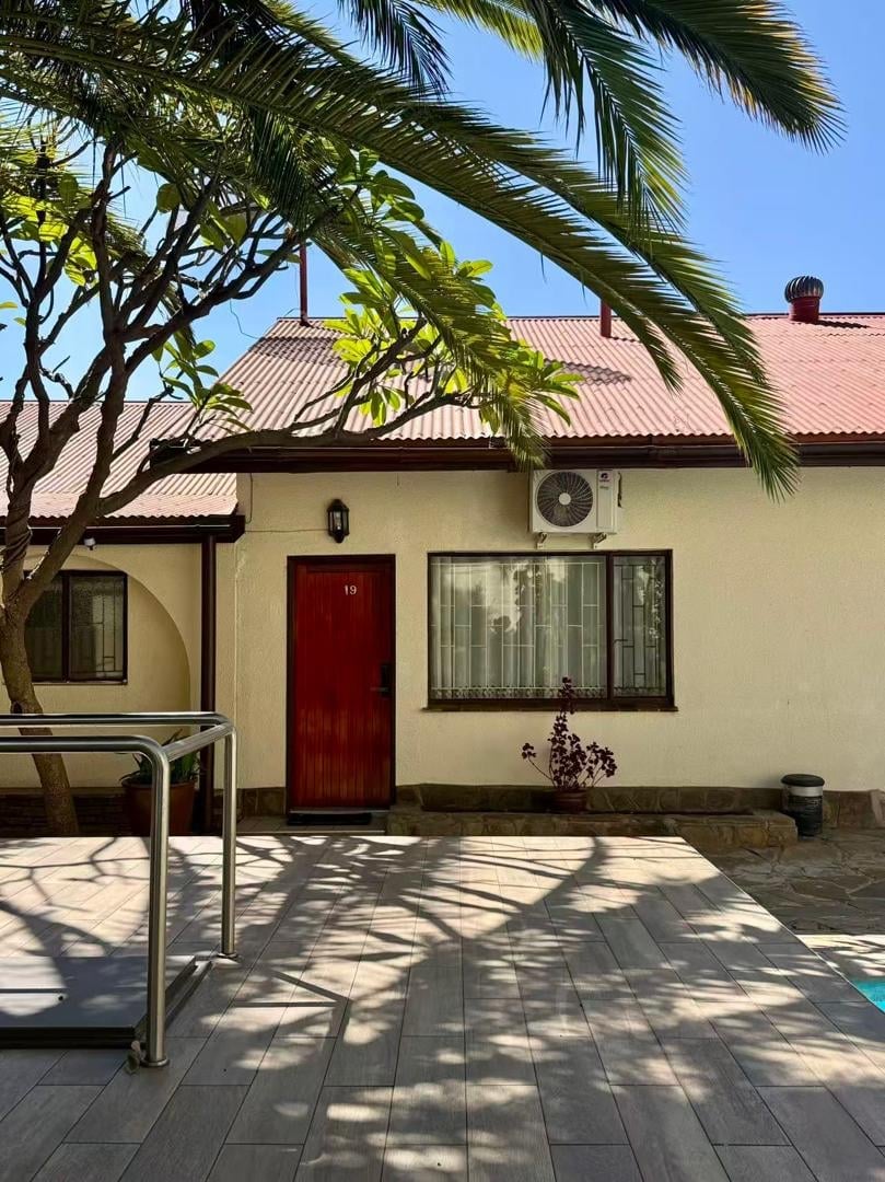 Cream-colored house with red door, metal roof, and palm trees, featuring a paved patio and lounge chair