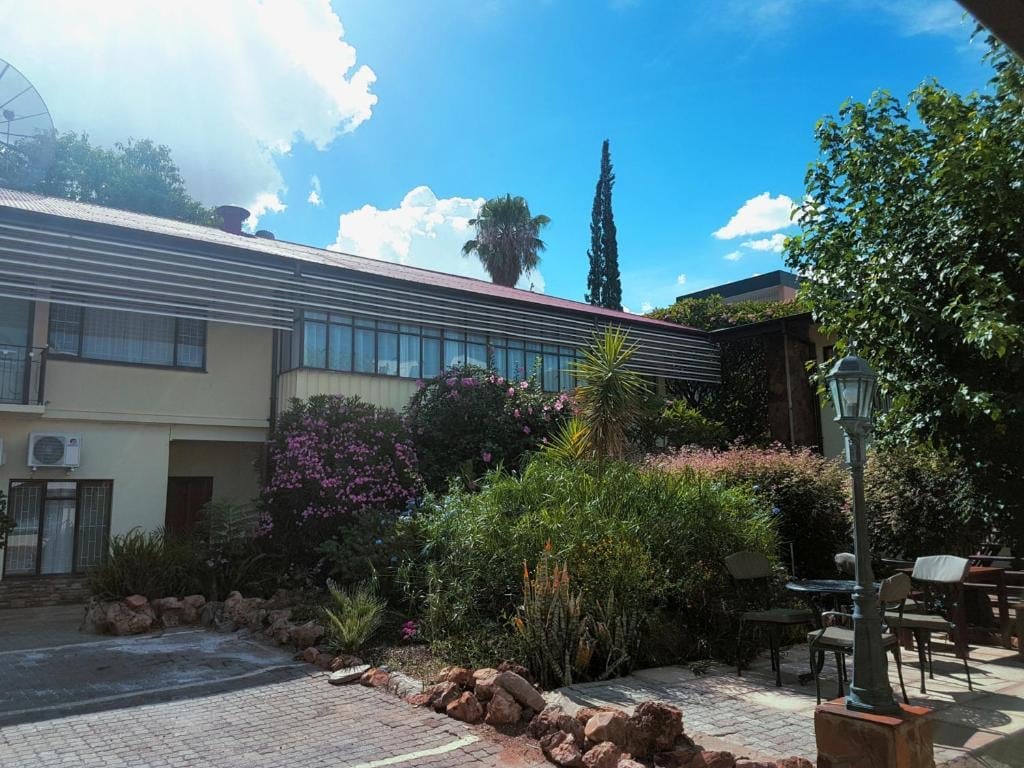 Modern single-story house with lush landscaping, including flowering shrubs and tall cypress tree, under blue sky with white clouds