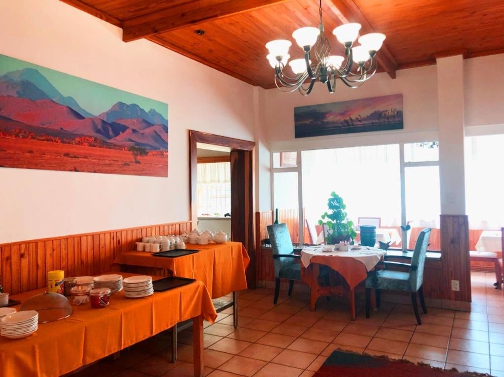 Dining room with wooden beams, orange linens, landscape artwork, large windows with mountain views, and modern chandelier