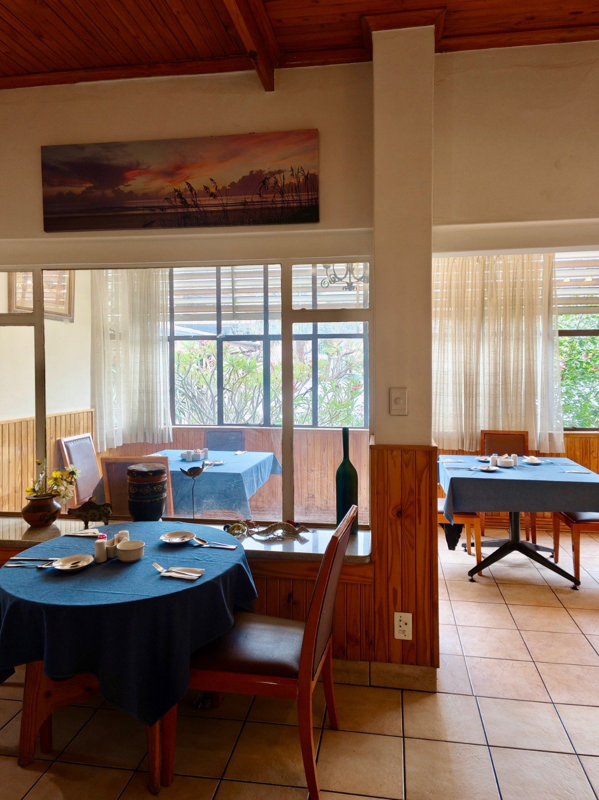 Modern dining room with round blue table, wooden furniture, large windows with sheer curtains, wood beam ceiling, and sunset artwork on wall