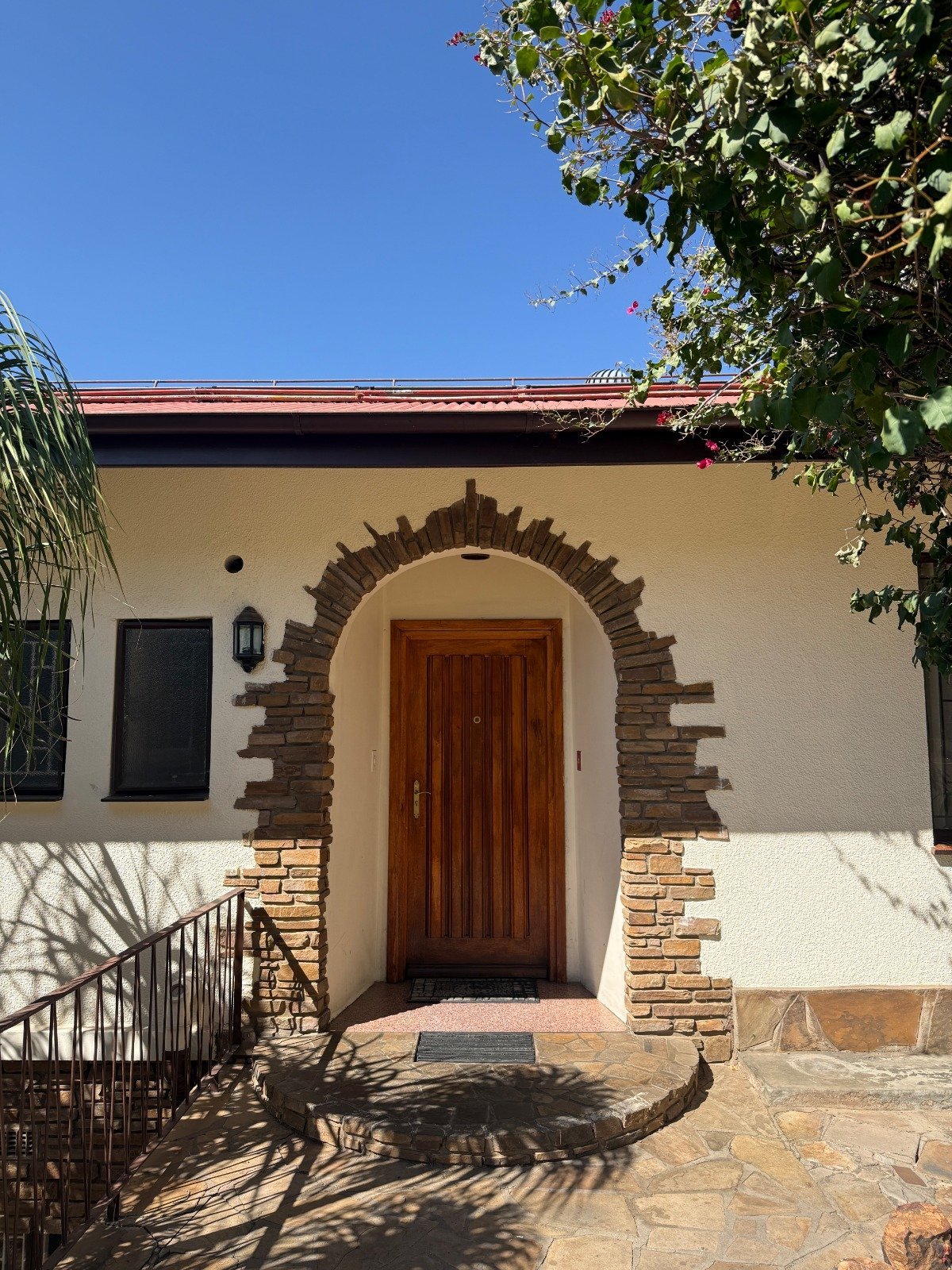 Mediterranean-style house entrance with white stucco walls, arched wooden door, brick details, and climbing ivy under blue sky