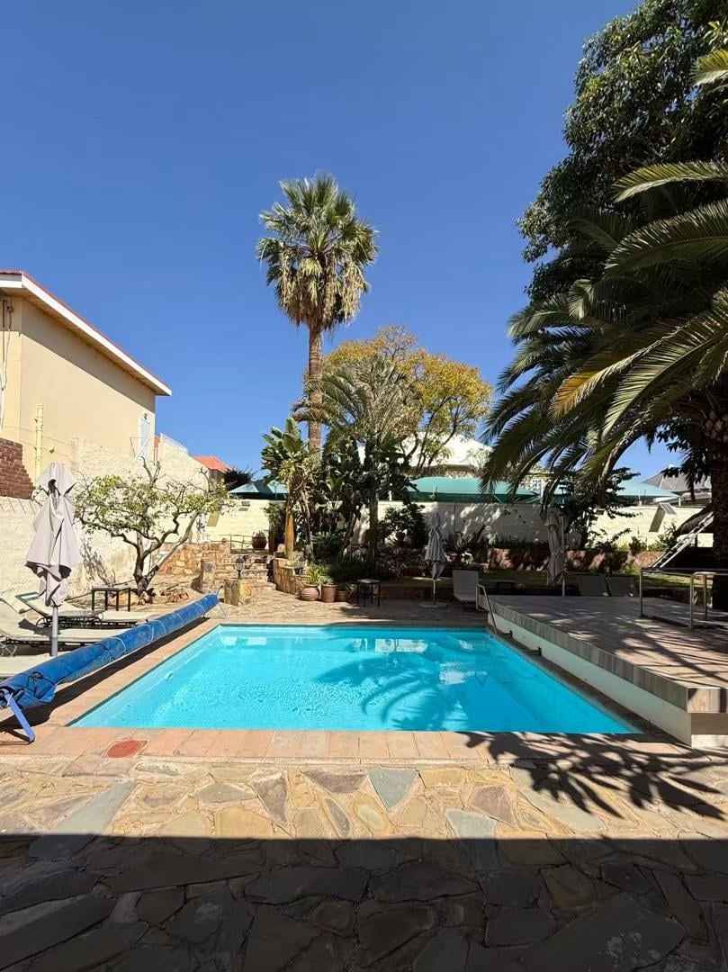 Bright turquoise swimming pool with stone patio, surrounded by palm trees and tropical vegetation, against a clear blue sky