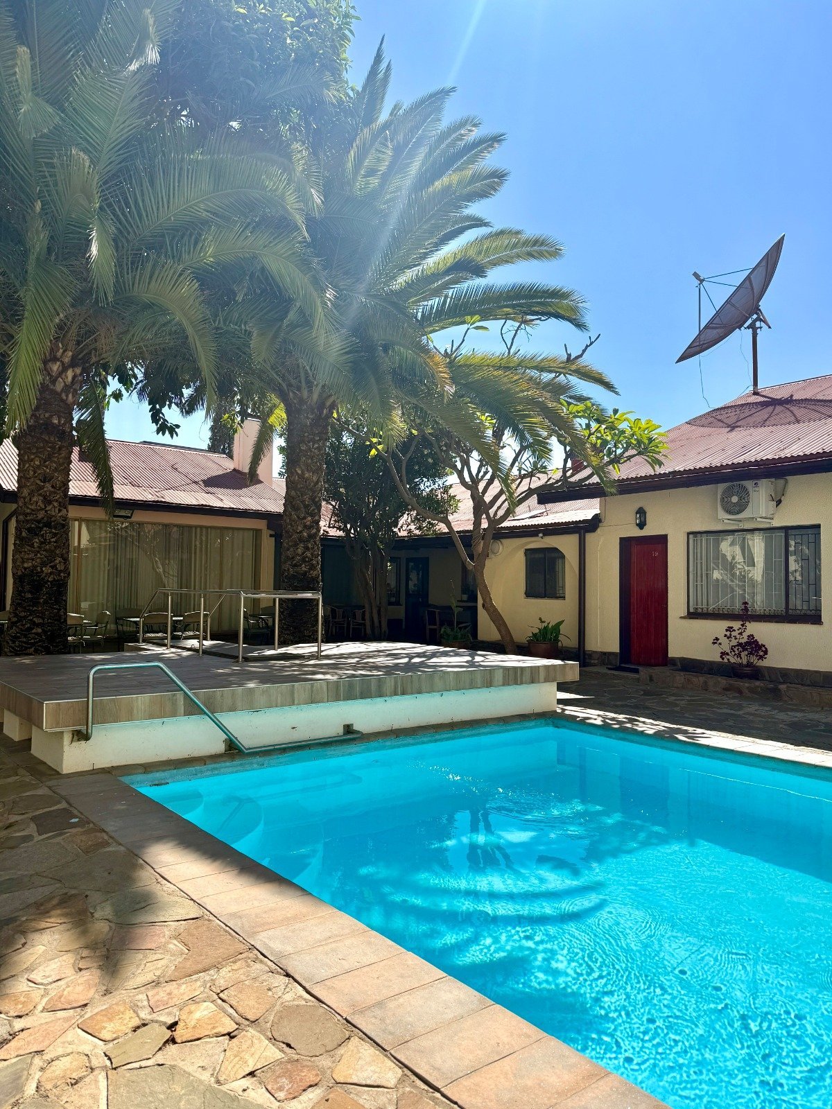 Backyard with bright blue swimming pool, spa tub, palm trees, cream-colored building with red door, and sunny clear sky