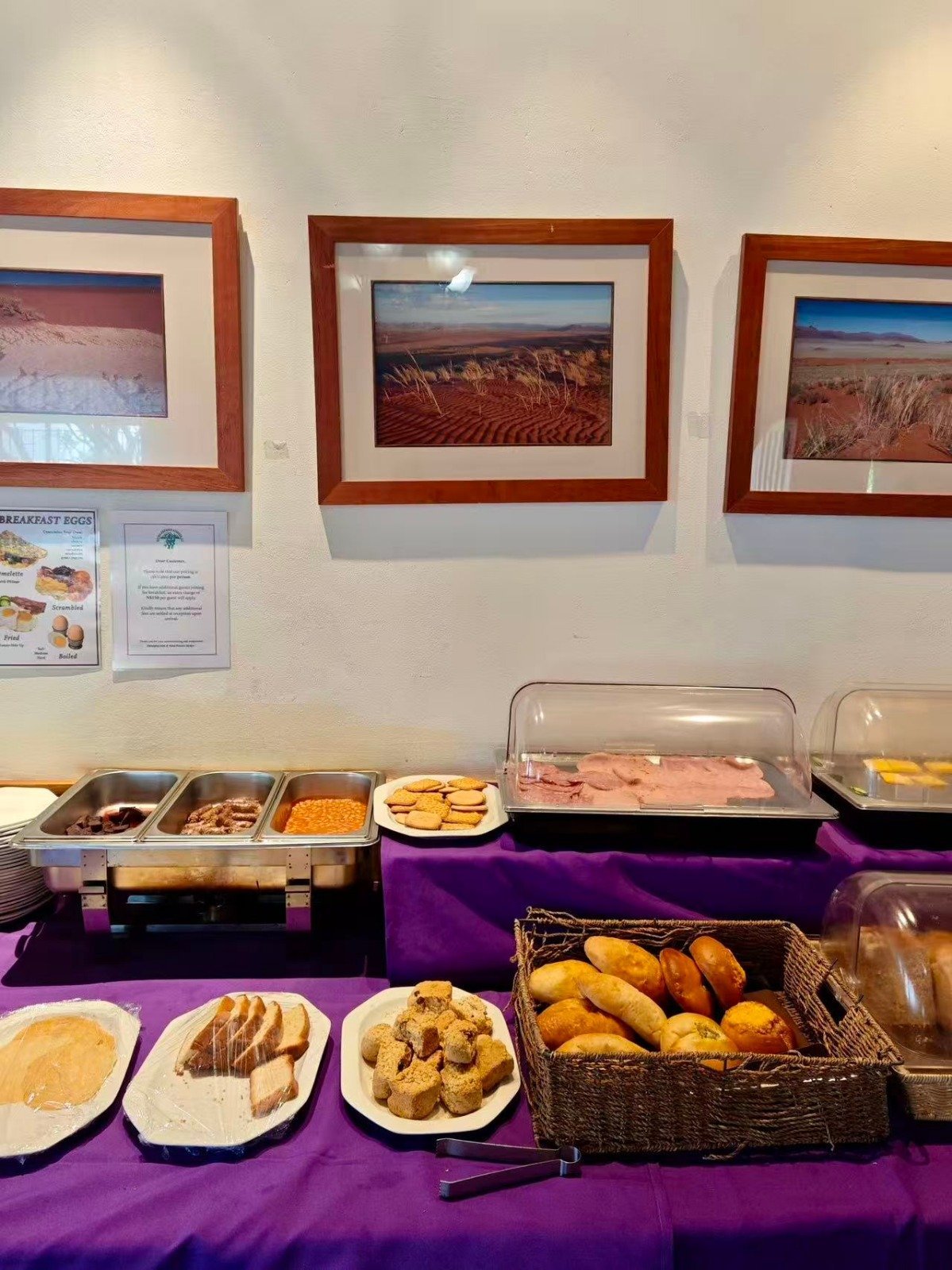 Breakfast buffet display with various pastries, breads, and hot food in metal trays on purple tablecloth, framed landscape photos on wall behind