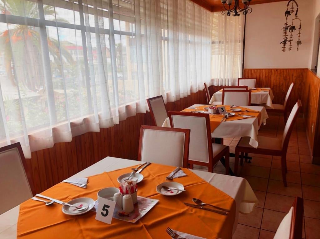 Elegant dining room with wooden tables set with orange cloths, place settings, and chairs by large windows with sheer curtains