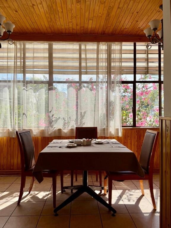 Dining room with round table and two chairs overlooking flower garden through large windows with sheer curtains and wooden ceiling