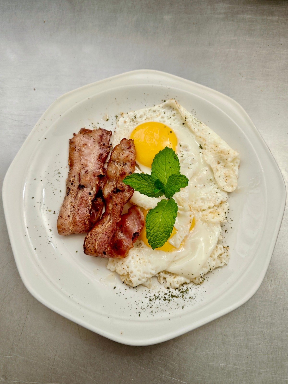 Breakfast plate with crispy bacon strips, fried egg with runny yolk, creamy mashed potatoes, fresh mint garnish, and black pepper on white plate