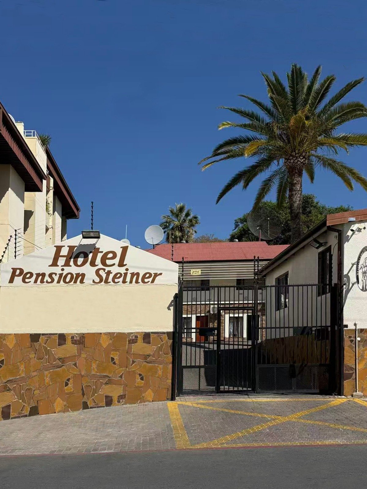 Front entrance of Hotel Pension Steiner with stone facade, metal gate, and palm tree against clear blue sky