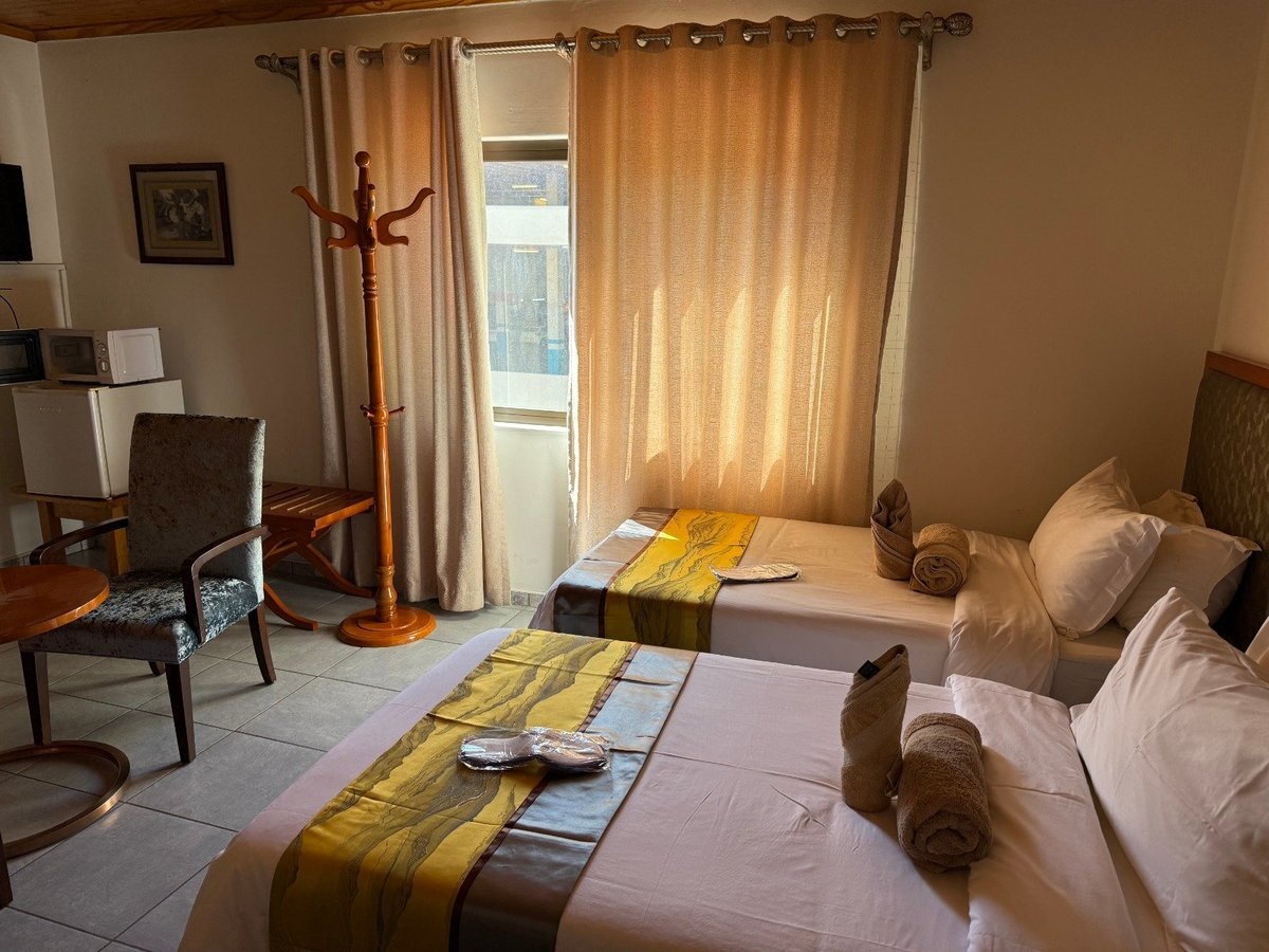 Warm-toned hotel room with two beds, golden curtains, wooden furnishings, and a seating area near the window.